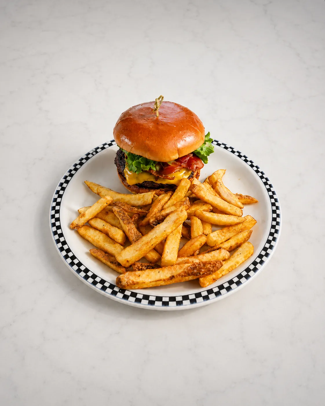 A bacon cheeseburger on a toasted brioche bun beside a pile of seasoned fries, on a black-and-white checkered rim plate set on white marble.